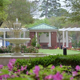 A serene garden scene featuring a fountain, vibrant flowers, and a gazebo in the background.