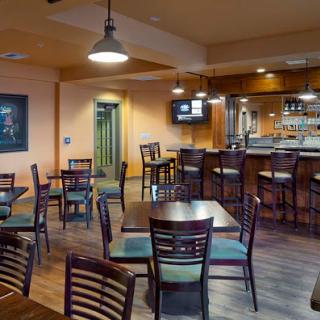A cozy restaurant interior featuring a dining area with wooden tables, chairs, and a bar in the background.