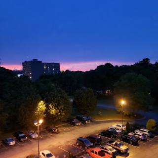 A serene evening view showcasing a vibrant sunset behind city buildings, accompanied by the glow of streetlights in a parking area.