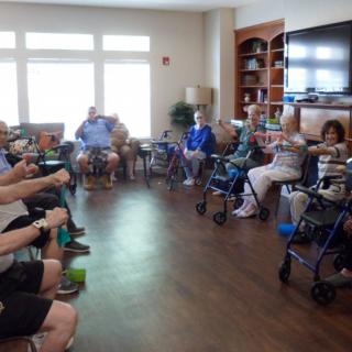A group of seniors participating in a seated exercise session inside a community room.