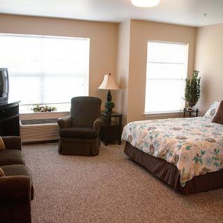 A cozy bedroom with a bed, armchairs, and a television, illuminated by natural light from large windows.