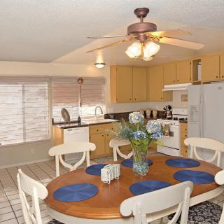 A cozy dining area with a wooden table set for four, featuring blue placemats and a centerpiece of flowers, adjacent to a kitchen equipped with modern appliances.