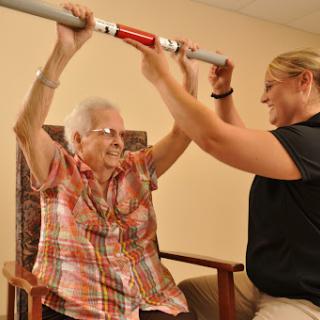 An elderly woman engages in an exercise session with the assistance of a caregiver, focusing on strength and mobility.