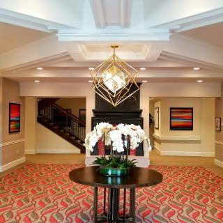 A modern hotel lobby with elegant decor, featuring a central table adorned with white flowers and a striking chandelier overhead.