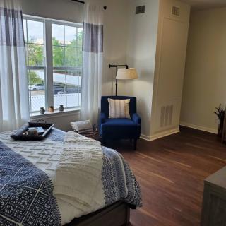 A cozy bedroom with a large window, featuring a bed with patterned bedding, a comfortable chair, and a standing lamp.