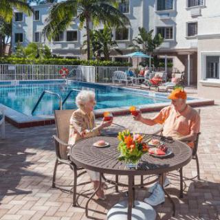 An elderly couple enjoys a meal and drinks by the pool in a tropical setting.