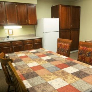A cozy kitchen with a dining table covered in a patterned tablecloth, surrounded by wooden chairs, featuring dark wooden cabinets and a white refrigerator.