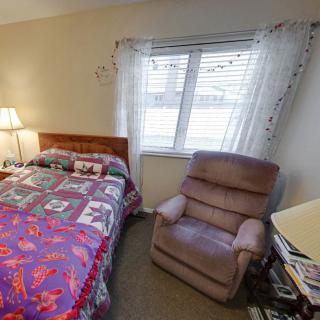 A cozy bedroom featuring a bed with a colorful quilt, a reclining chair, a window with curtains, and a lamp.