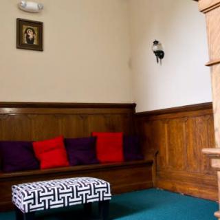 A cozy interior room with wooden paneling, bench seating adorned with colorful cushions, and a patterned ottoman. A portrait and wall sconce decorate the walls.