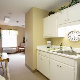 A small kitchen area with white cabinets and a sink, featuring a wall clock and natural light coming through the window.