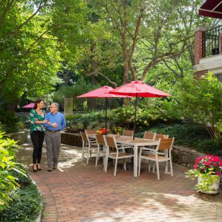A couple walking in a lush garden with patio furniture and red umbrellas.