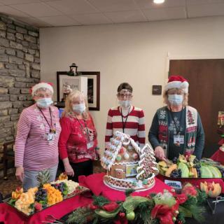 Four people in festive attire stand behind a table with a decorated gingerbread house and holiday decorations.