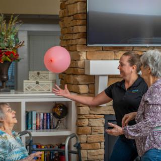 A caregiver engages with seniors in a lively indoor activity, tossing a pink balloon to promote fun and interaction.