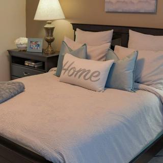 A cozy and well-decorated bedroom featuring a neatly made bed with multiple pillows and a 'Home' accent pillow, alongside a bedside lamp.