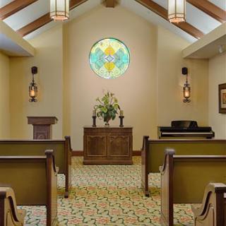 A serene chapel interior featuring wooden pews, a floral arrangement at the altar, and beautiful stained glass.