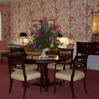 A traditionally decorated dining room with a classic wooden table and chairs, featuring a floral centerpiece and patterned wallpaper.