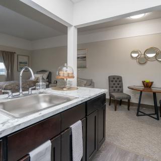 A modern apartment interior featuring a kitchen area with a sink and countertop, leading into a living space with chairs and a table.