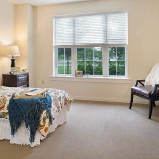 A cozy bedroom with a large window, neatly arranged furniture including a bed, lamp, and chair.