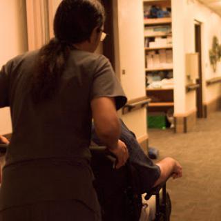 A nurse assists a patient in a wheelchair down a hospital corridor.