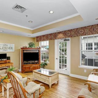 A cozy living room with a television, comfortable seating, and natural light from large windows.