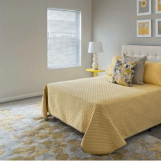 A modern bedroom with yellow bedding and decorative pillows, featuring a lamp and framed artwork on the neutral-colored walls.