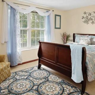 A cozy bedroom featuring a wooden bed with light blue bedding, a patterned rug, and large windows with sheer curtains.
