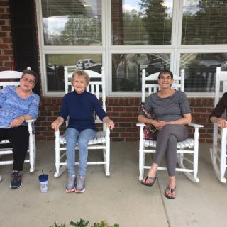 A group of people sitting on white rocking chairs on a porch.
