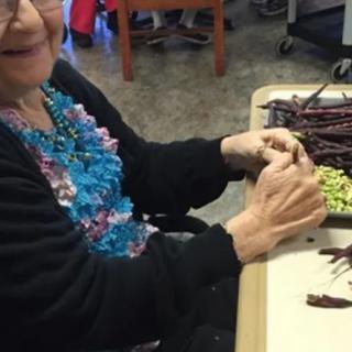 Elderly woman sitting at a table, preparing vegetables with a smile.