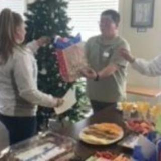 People exchanging gifts at a holiday party with food on the table and a decorated Christmas tree in the background.