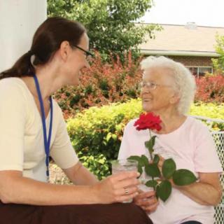 A caregiver engaging in a friendly conversation with an elderly woman holding a flower in a garden setting.