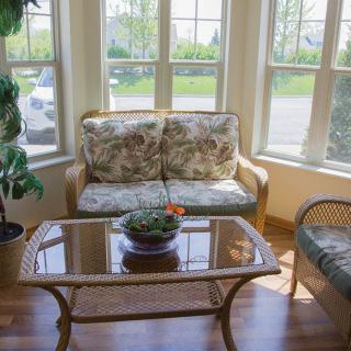 A cozy sunlit living room with a floral sofa, glass-top coffee table, and large windows.