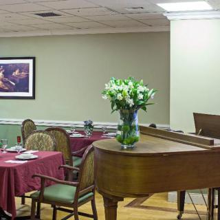 A cozy dining area featuring a piano and tables with red tablecloths, adorned with a vase of flowers.