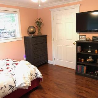 A cozy bedroom with warm peach walls, featuring a bed with floral bedding, a tall dresser, and a wall-mounted television above a small bookshelf.