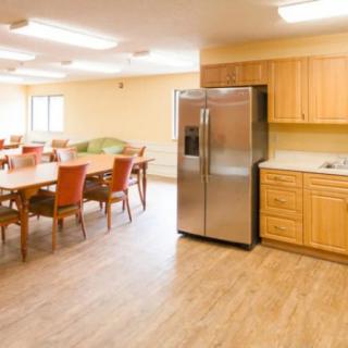 A spacious kitchen and dining area with wooden flooring, featuring a large refrigerator, wooden cabinets, and multiple tables and chairs.