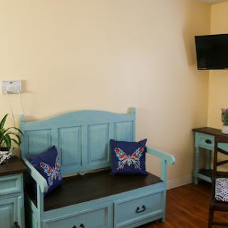A cozy living room space featuring a light blue bench with decorative pillows, a small plant, and a television mounted on the wall.