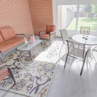 A cozy patio area with comfortable seating, a glass table, and decorative rugs, illuminated by natural sunlight.