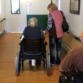 A caregiver helps an elderly person in a wheelchair navigate down a hallway.