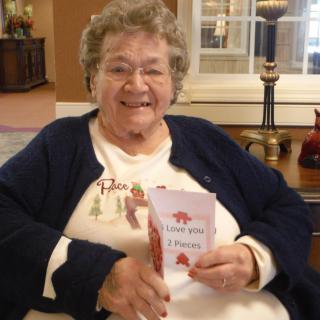 An elderly woman sitting indoors, smiling and holding a book.