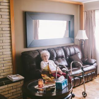 An elderly woman sitting on a sofa in a cozy living room with a walker nearby.