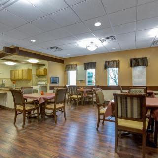 A spacious dining area with wooden flooring, featuring several tables and chairs, adjacent to a kitchen space.