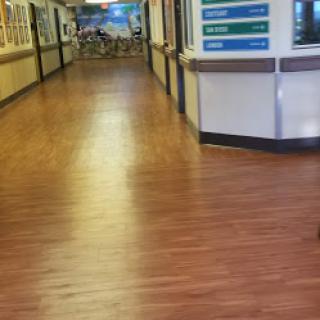 A view of a hospital hallway with wooden flooring, featuring multiple doors and directional signs.