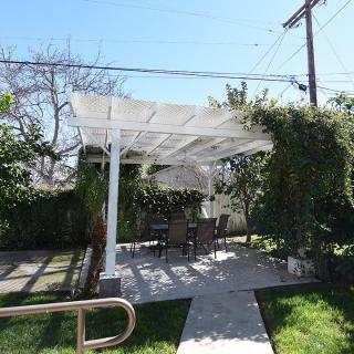 A serene outdoor patio with a white pergola providing shade, surrounded by lush greenery.