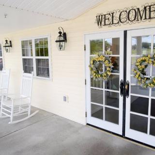 A cozy porch entrance with a welcome sign and decorative wreaths on the glass doors, featuring a white rocking chair and outdoor lighting.