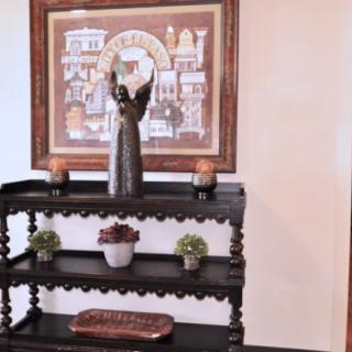 A decorative shelf with a statue, candles, and small plants, set against a wall featuring framed artwork.