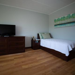 A minimalist bedroom featuring a single bed, a wooden dresser with a television on top, and light wood flooring.