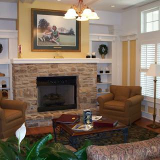A cozy living room featuring a stone fireplace, two armchairs, and a coffee table, with a painting above the mantelpiece.