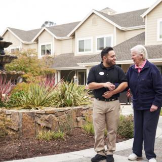 A caregiver assisting a senior citizen as they walk outdoors in a landscaped area.