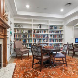 A cozy library room featuring a fireplace, bookshelves filled with books, a table with chairs, and a computer station.