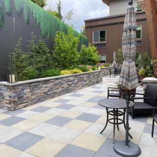 A modern patio area with tables, chairs, and umbrellas surrounded by lush greenery and a stone wall.