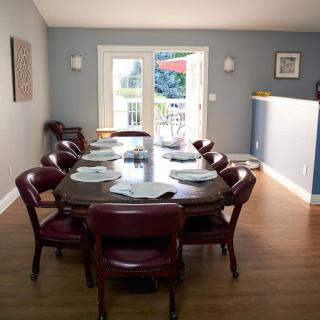 A dining room featuring a long table with place settings, surrounded by chairs and illuminated by natural light from large windows.